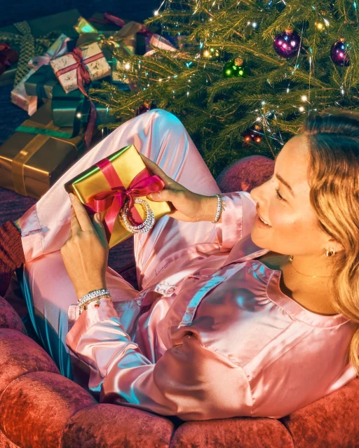 A woman in pink pajamas sits near a decorated Christmas tree, smiling as she looks at a gift wrapped in gold with a pink bow.