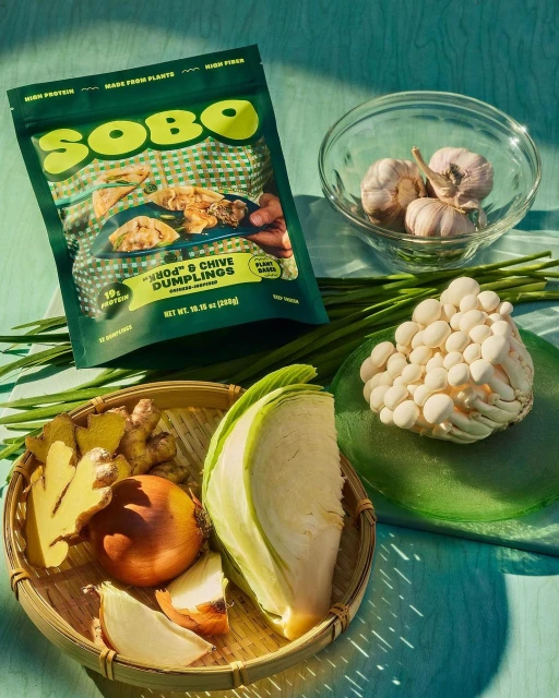 SOBO dumpling package with ingredients like onion, garlic, cabbage, mushrooms, and ginger on a green surface in bright light.