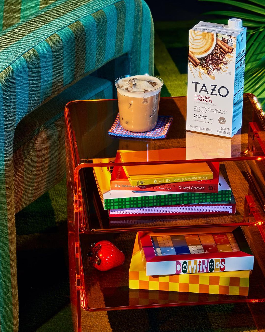 TAZO iced chai latte carton and glass on a mirrored cart with books and a red strawberry prop, teal chair in the background.