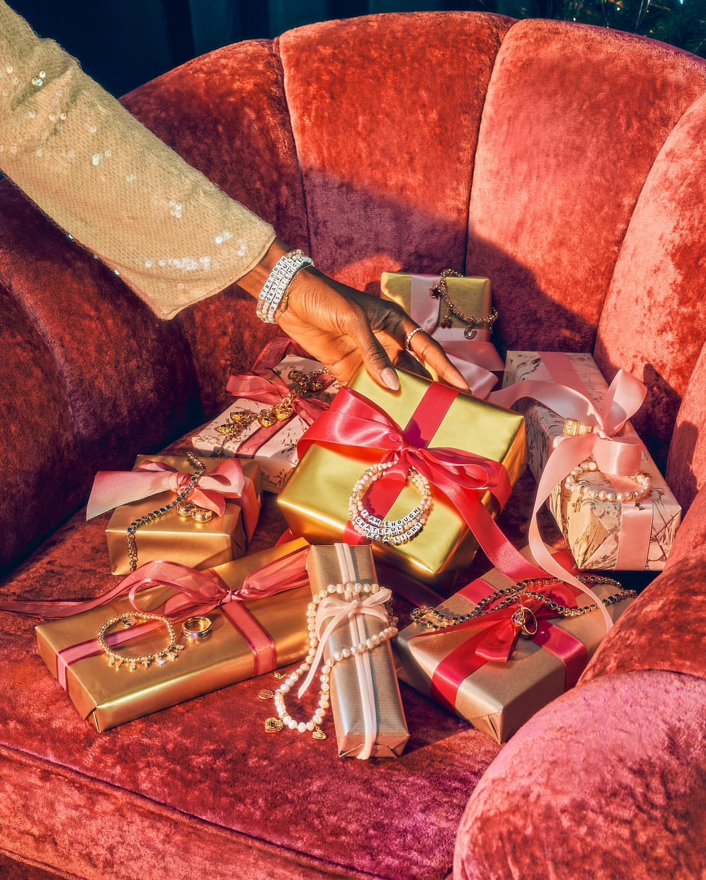 Gifts wrapped in gold paper and tied with pink ribbon are displayed on a red velvet chair, while an arm wearing a sequin sleeve reaches in.