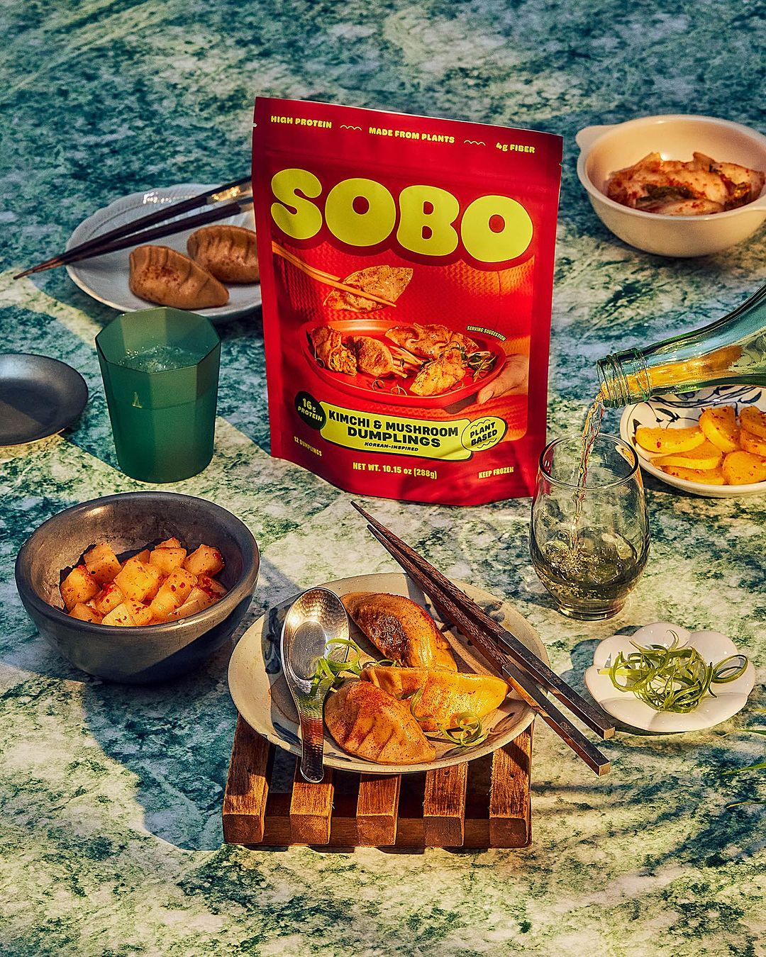 SOBO kimchi and mushroom dumplings arranged on a green marble surface with bowls of sides and drinks.