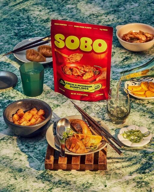 SOBO kimchi and mushroom dumplings arranged on a green marble surface with bowls of sides and drinks.