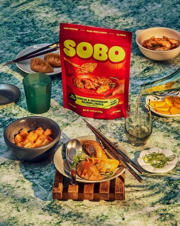 SOBO kimchi and mushroom dumplings arranged on a green marble surface with bowls of sides and drinks.