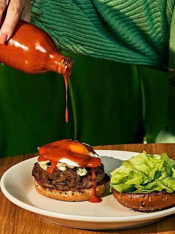 A partially eaten burger sits on a white plate on a wooden table with brown sauce being poured on it, with a green sweater sleeve.
