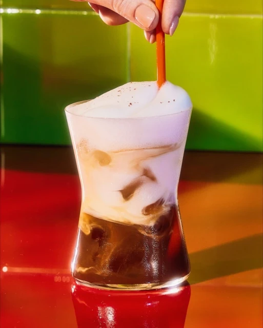 Iced latte in a clear glass with orange straw, set against a vibrant red and green background, and woman's hand visible.