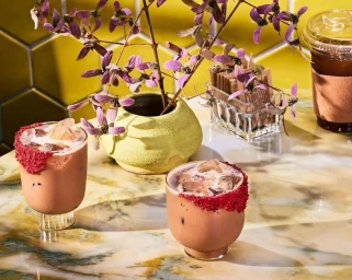 Two iced coffee drinks in ribbed glasses with red rims on a marble table with a yellow vase and purple flowers by Jenny Huang.