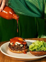 Hot sauce pouring from a glass bottle onto a gourmet burger on a white plate by photographer Jenny Huang
