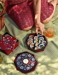 Overhead view of a woman seated on a green plaid picnic blanket reaching for sushi rolls from decorative lacquer boxes