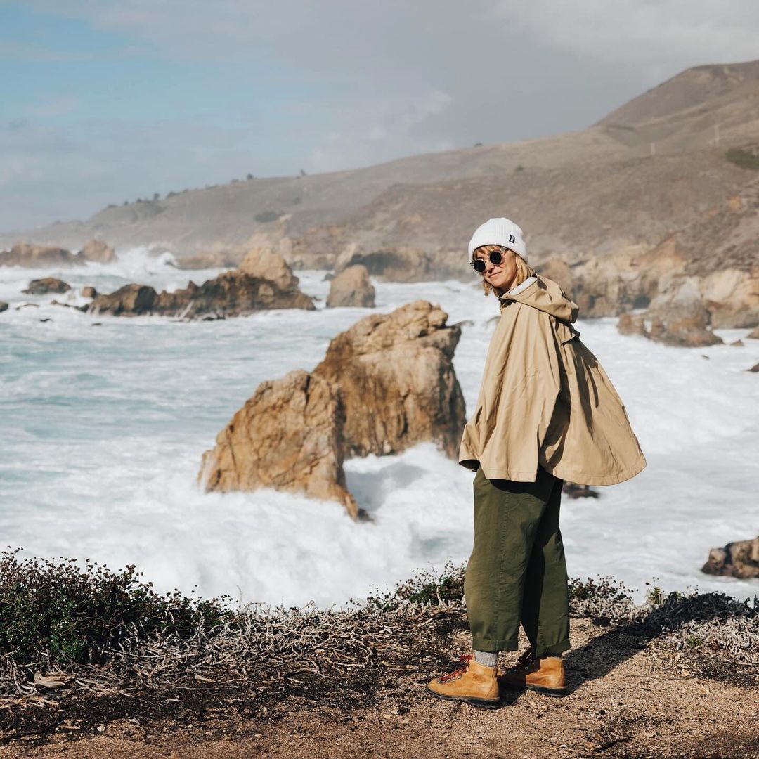 Woman in beanie and poncho standing on coastal cliff with ocean waves crashing against rocks on a sunny day in a landscape orientation.