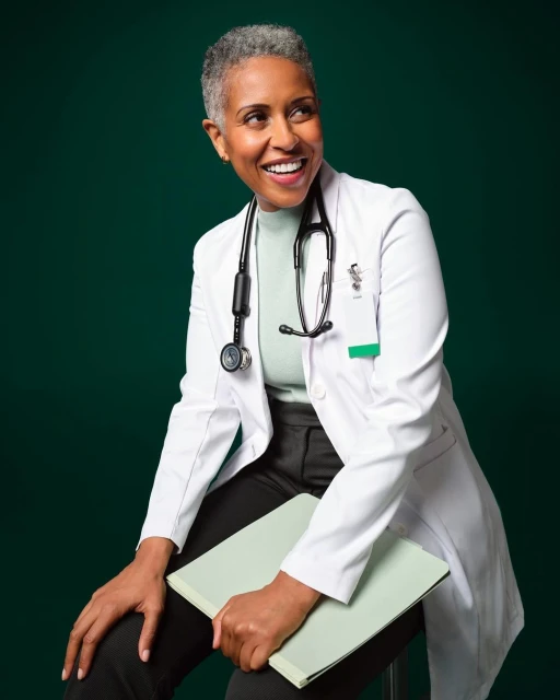 Smiling African-American doctor in a white coat sits against a dark green background, holding a light green folder and wearing a stethoscope.