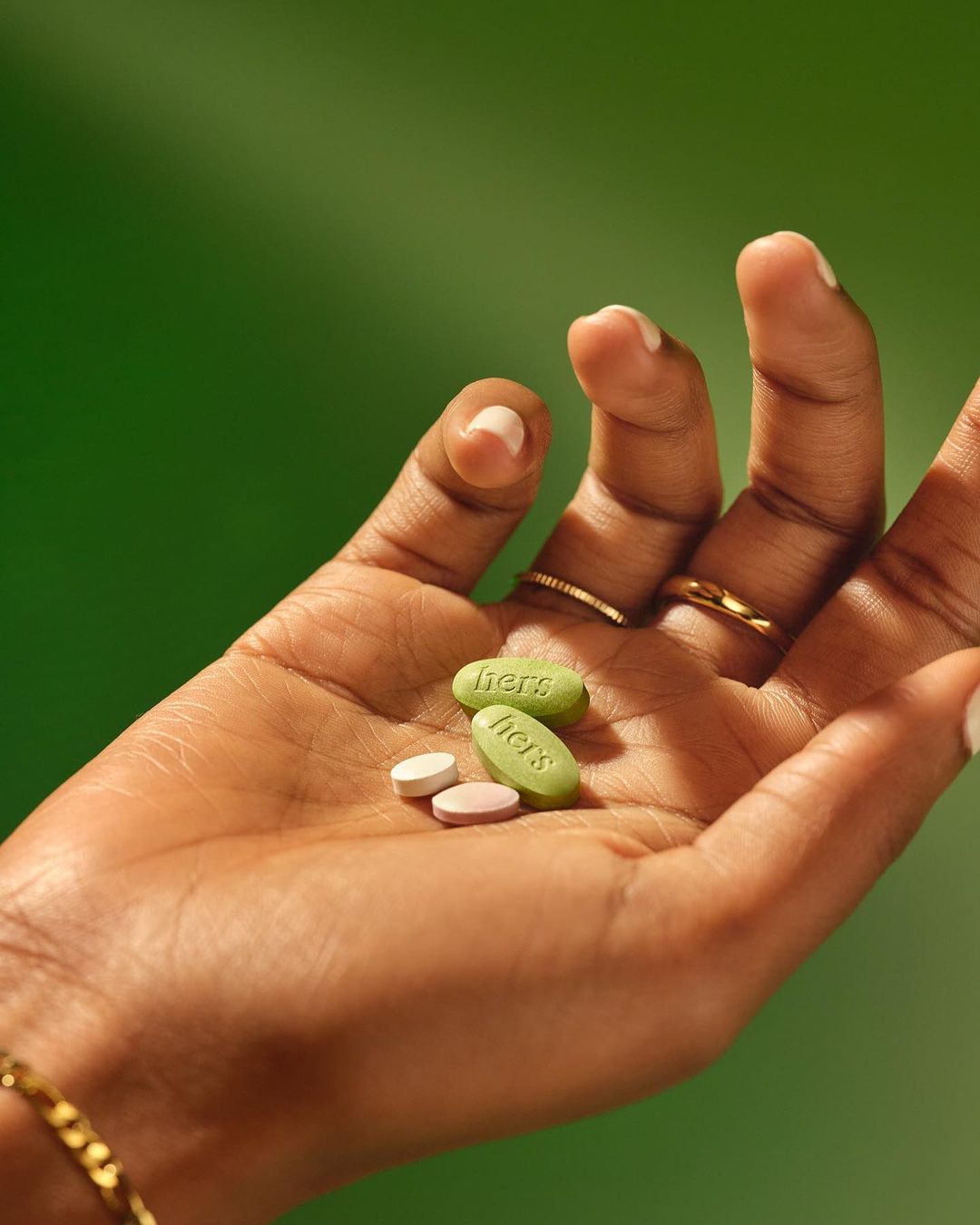 Close-up of a dark-skinned hand holding three capsules with the word 'hers' printed on them, set against a solid green background.