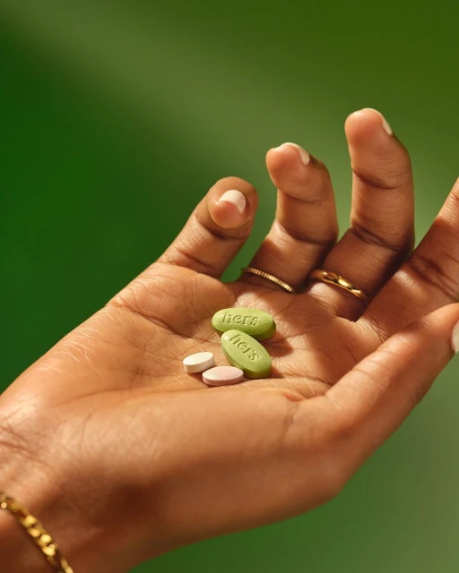 Close-up of a dark-skinned hand holding three capsules with the word 'hers' printed on them, set against a solid green background.