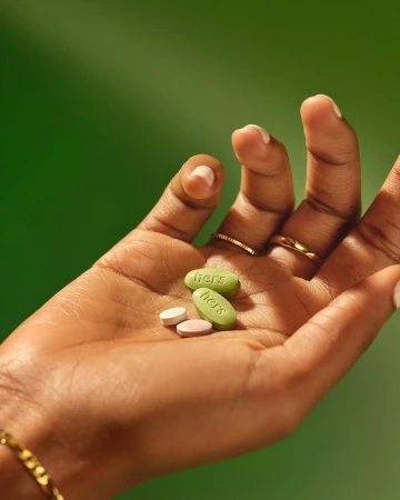 Close-up of a dark-skinned hand holding three capsules with the word 'hers' printed on them, set against a solid green background.