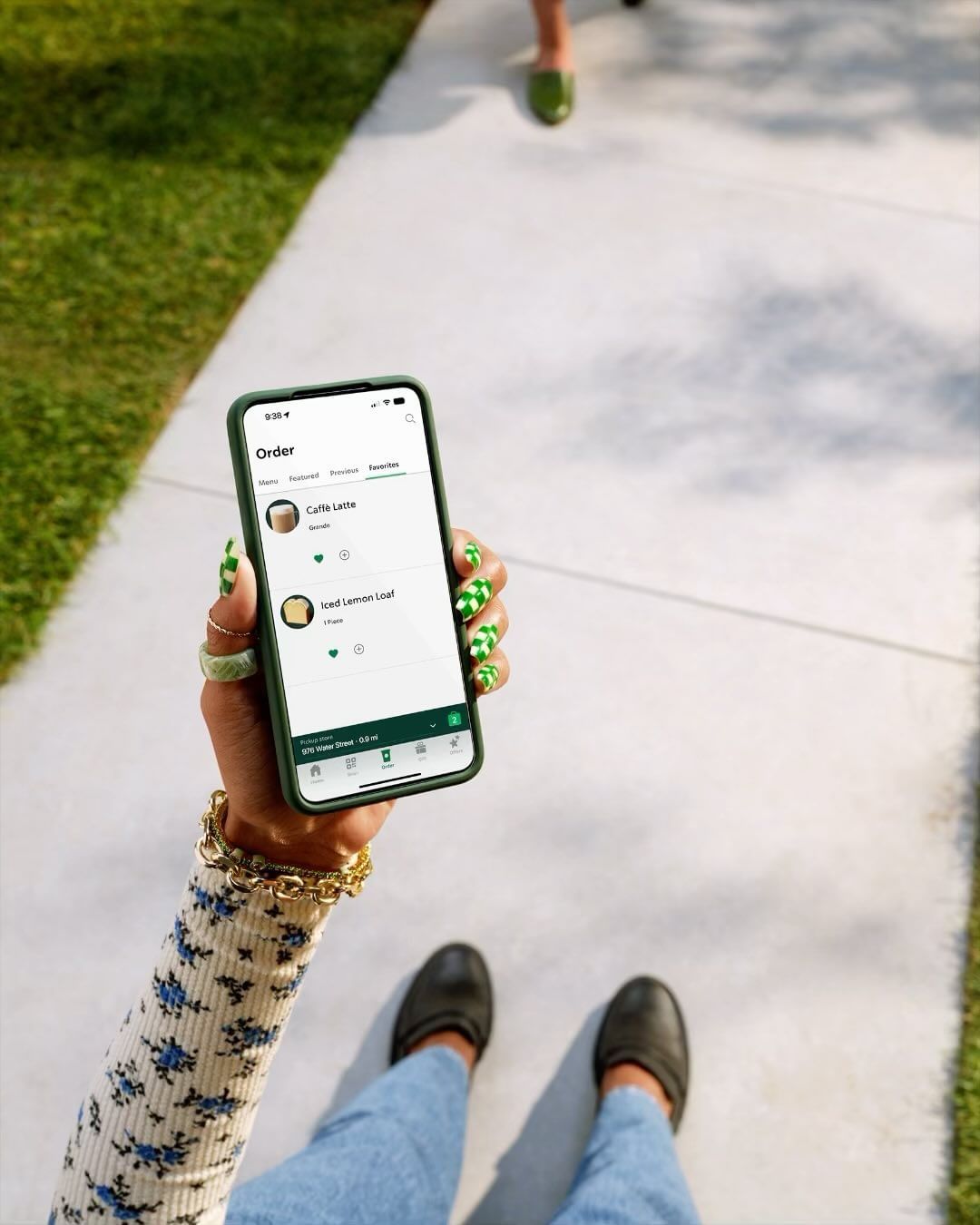 Person holding a phone showing a coffee order app with green patterned nails, walking on a sidewalk with green grass alongside.