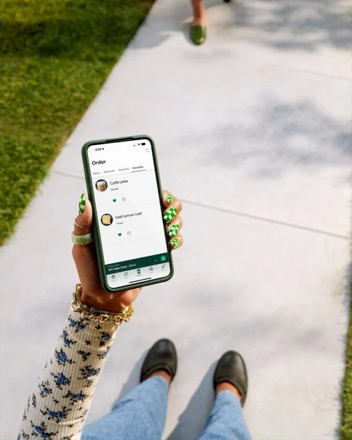 Person holding a phone showing a coffee order app with green patterned nails, walking on a sidewalk with green grass alongside.