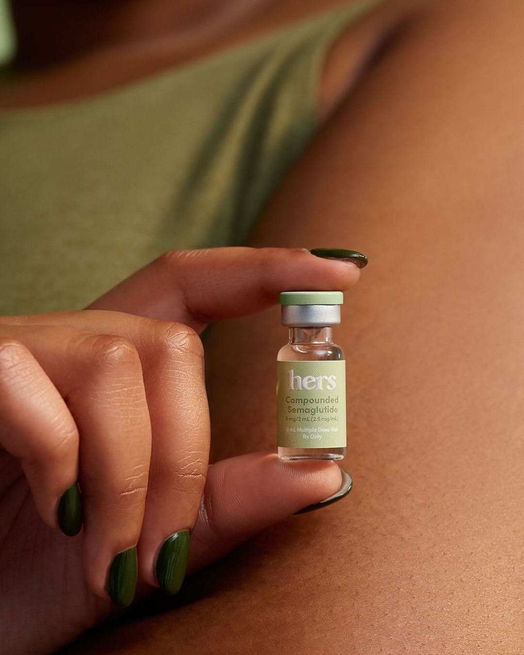 Adult female hand holding Hers compounded semaglutide vial. Close-up of dark olive manicure, light olive tank top, neutral tan skin.