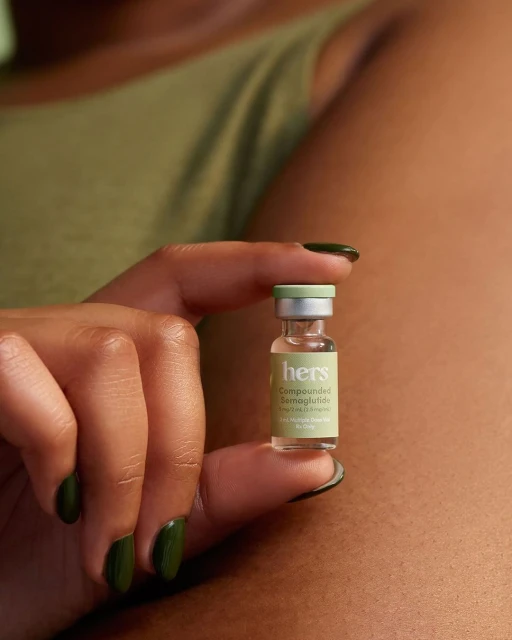 Adult female hand holding Hers compounded semaglutide vial. Close-up of dark olive manicure, light olive tank top, neutral tan skin.