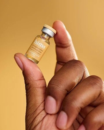 Close-up of dark-skinned hand holding small bottle of semaglutide against a yellow background, showcasing skincare or medication.