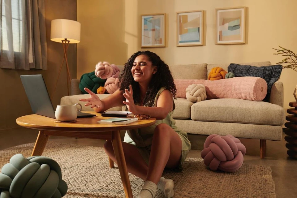 Smiling woman video conferencing on a laptop indoors, seated on a rug near a wooden table, sofa, and knot-shaped cushions.