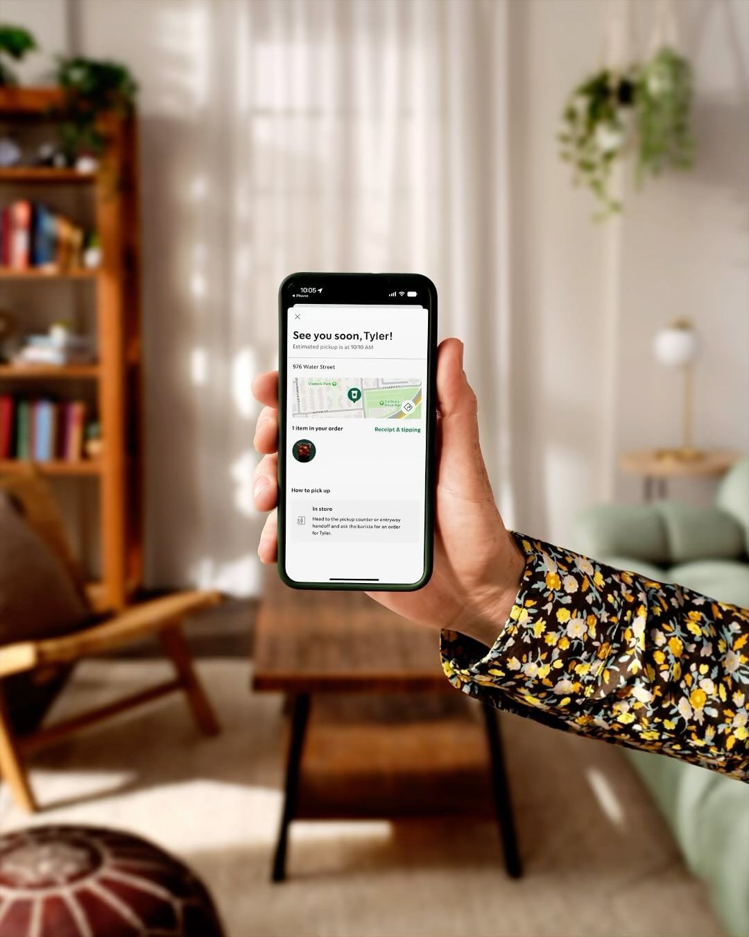 Person holds a smartphone displaying an order tracking screen, inside a living space with a table and shelving unit in view.