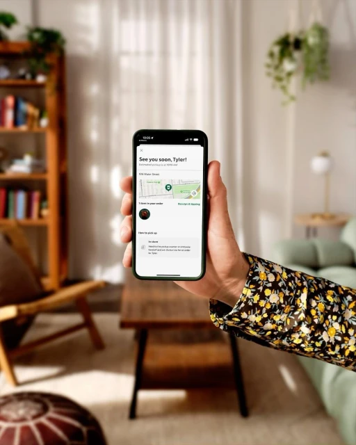 Person holds a smartphone displaying an order tracking screen, inside a living space with a table and shelving unit in view.