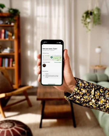 Person holds a smartphone displaying an order tracking screen, inside a living space with a table and shelving unit in view.