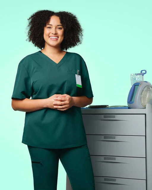 Smiling female healthcare worker in green scrubs stands in a studio setting with medical equipment. Focus on medical professionals.