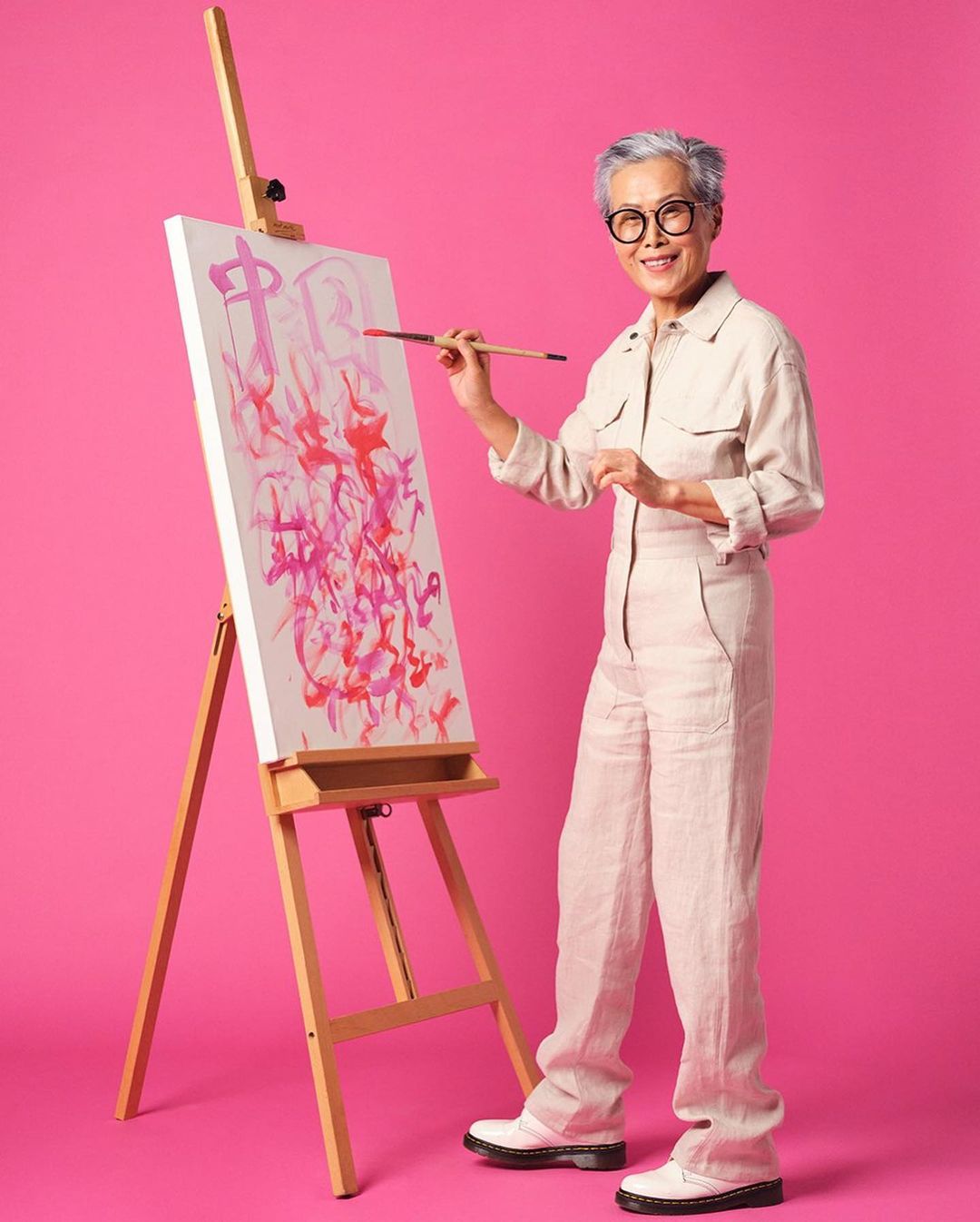 An elderly Asian woman in a jumpsuit stands indoors smiling, painting an abstract design on a canvas on a wooden easel.