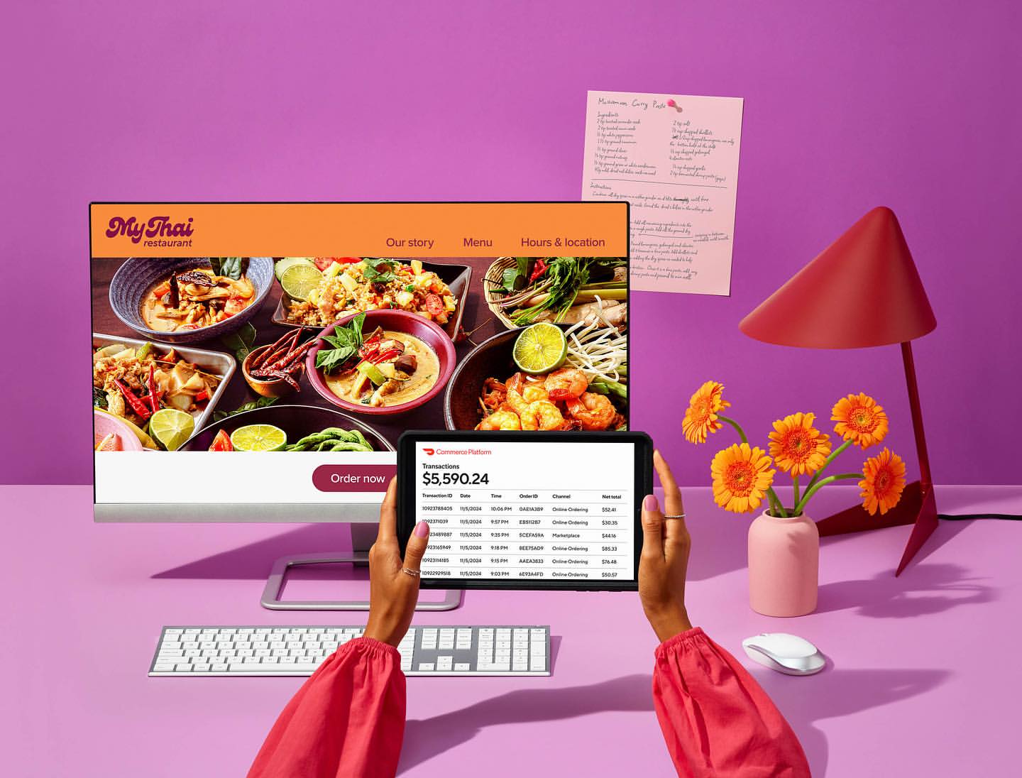 Close-up of a purple desk featuring a monitor displaying a Thai restaurant website, a tablet with transaction details, and floral arrangement.