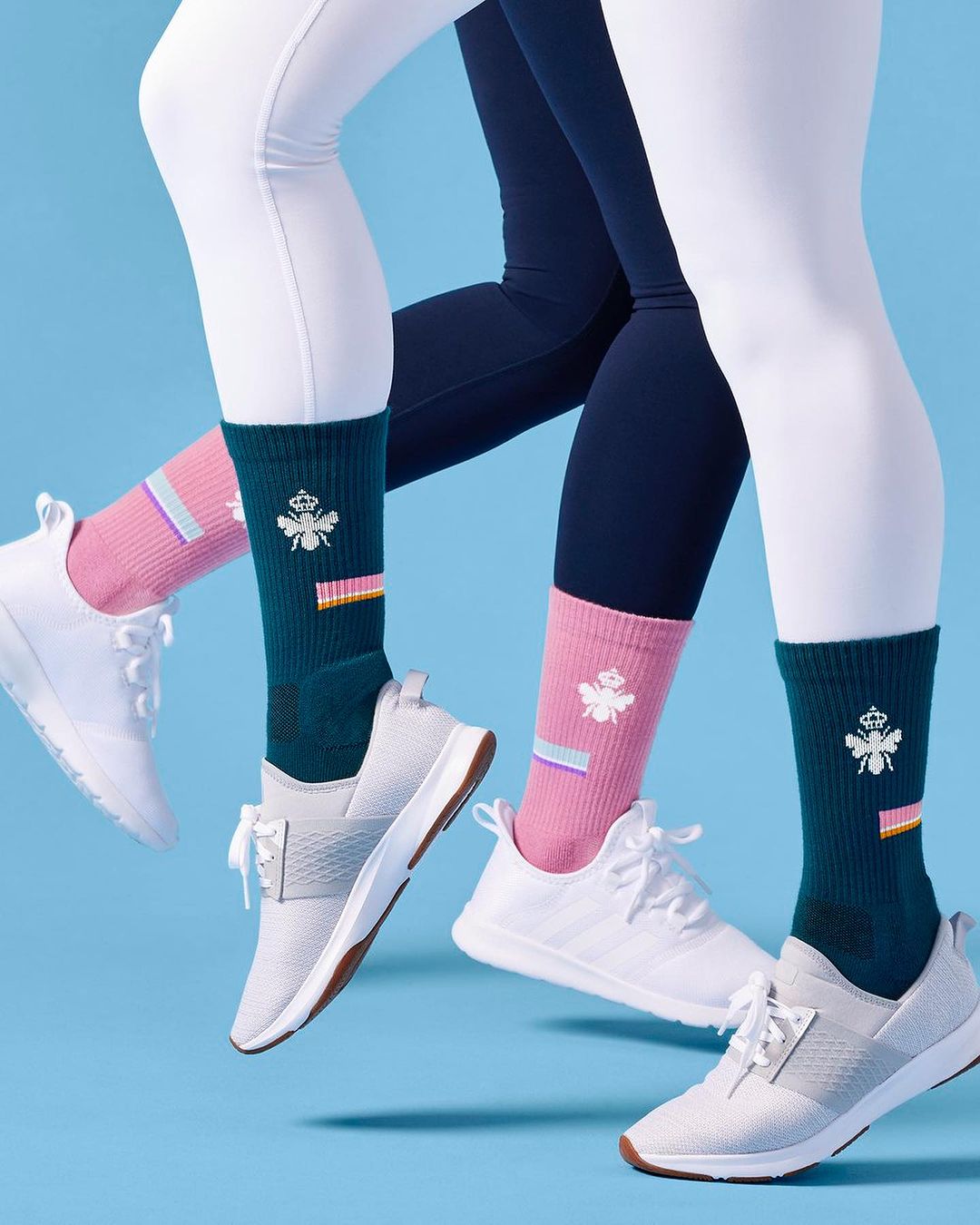 Two models showcase athletic socks with white sneakers against a light blue background in a studio fashion product shot.