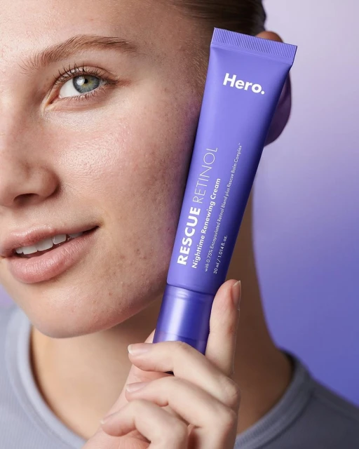 Close-up of a fair-skinned woman holding a tube of Hero Rescue Retinol Nighttime Renewing Cream against a lavender background gradient.