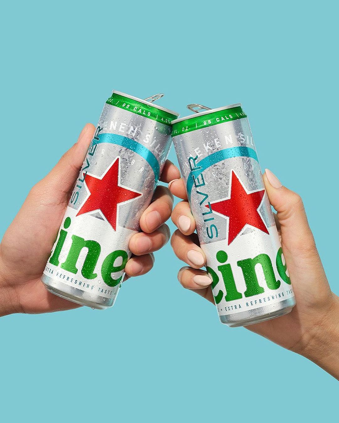 Two hands toasting with silver Heineken Silver beer cans against a blue background, with focus on the cans and fingers.