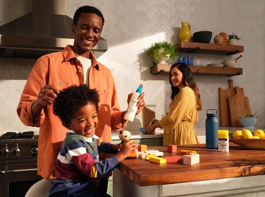 A smiling father sprays a toddler's hair inside a well-lit kitchen as a woman washes dishes at the sink in the background.
