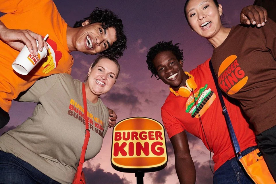 Four smiling young adults of diverse ethnicities pose in Burger King branded t-shirts against a cloudy sky with Burger King sign.
