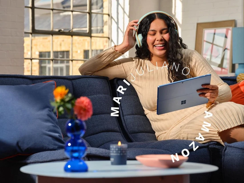 A woman sits on a blue couch using a tablet and wearing headphones, with a blue glass vase and candle on a nearby table.