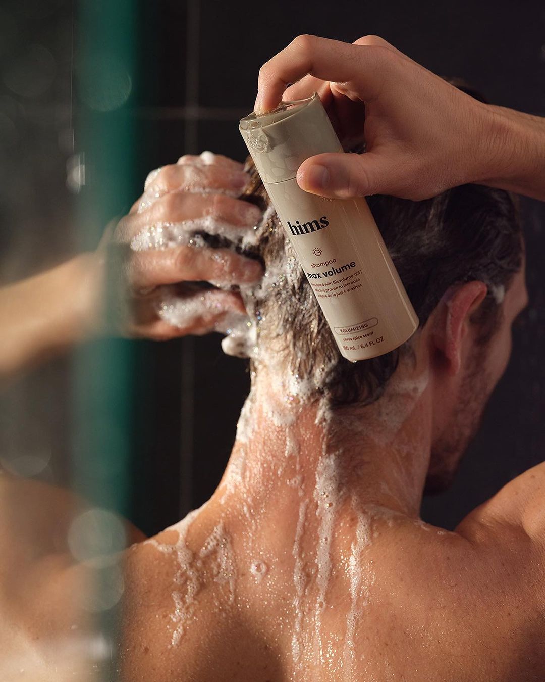 Adult male washing hair in shower with foaming shampoo, holding bottle of hair product in hand in a dimly lit bathroom setting.