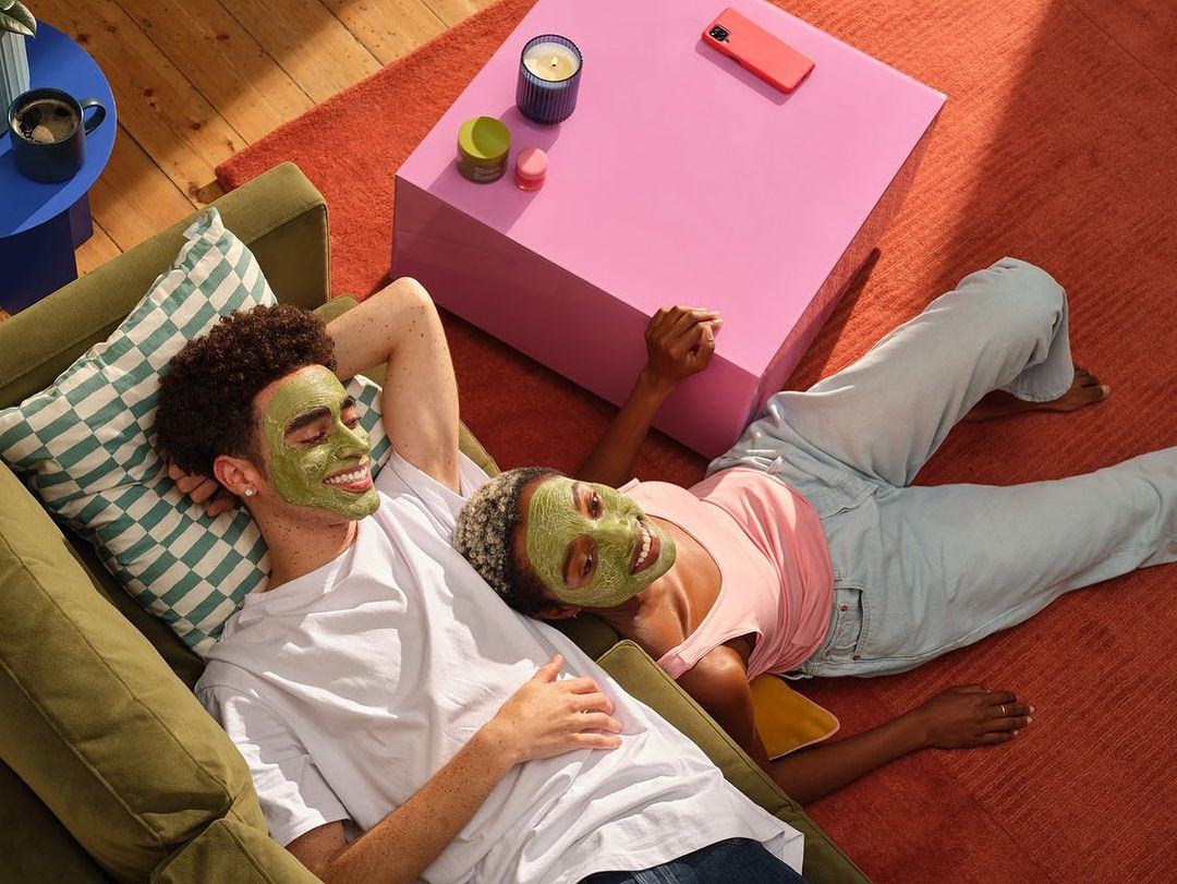 A smiling couple with green face masks relaxing on an olive sofa in a brightly lit room with a pink table and orange rug.