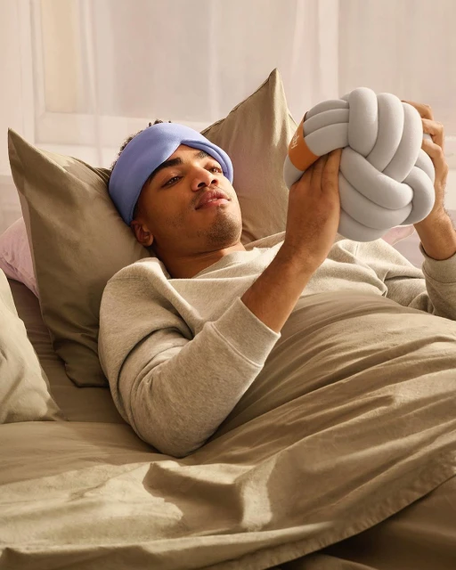 Man with headband rests in bed holding a knot pillow, showcasing a cozy and comfortable scene with soft lighting and neutral color palette.