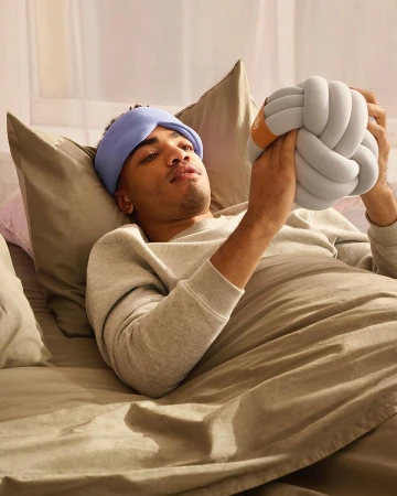 Man with headband rests in bed holding a knot pillow, showcasing a cozy and comfortable scene with soft lighting and neutral color palette.