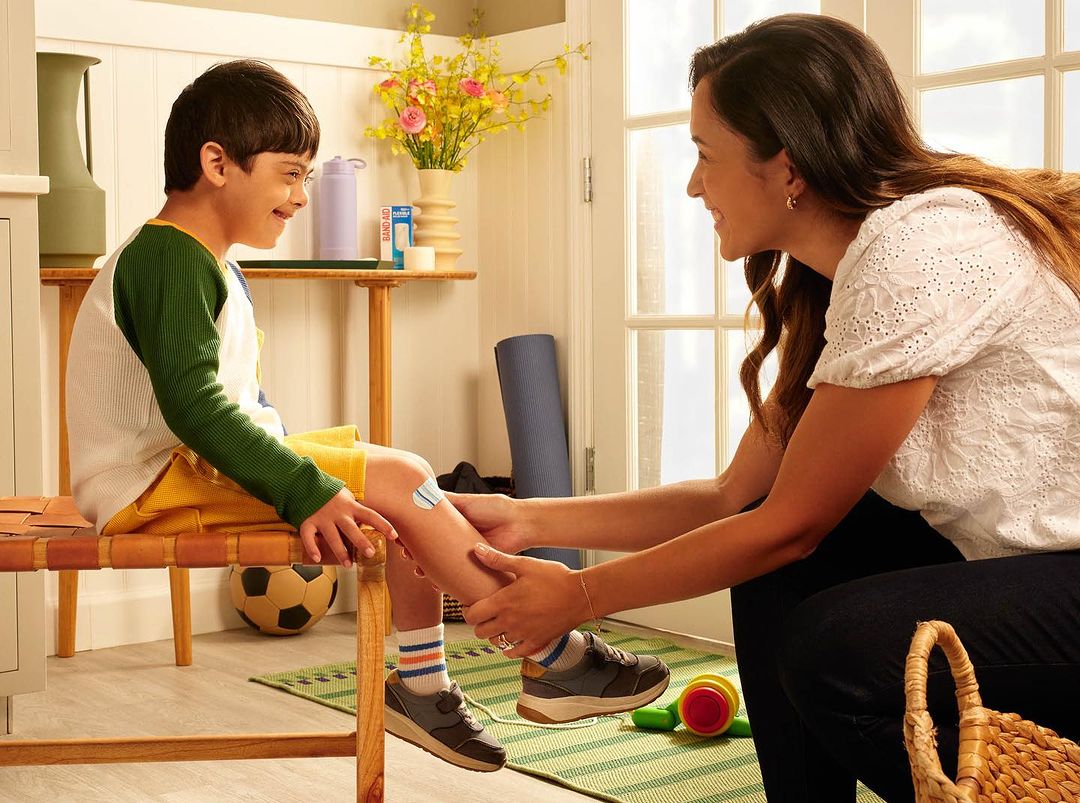 Smiling boy with patch on his leg seated on a bench as a woman examines his leg in a bright indoor setting, showcasing healthcare.