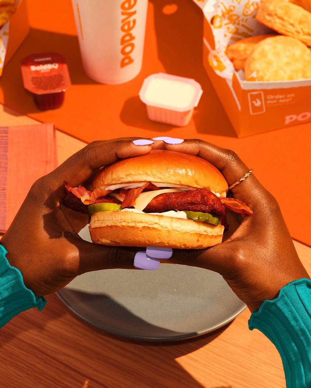 Overhead shot of person holding a sandwich with dark skin against an orange backdrop featuring Popeyes cup, box, and sauce.