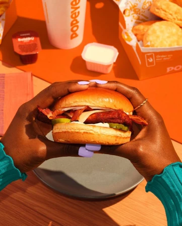 Overhead shot of person holding a sandwich with dark skin against an orange backdrop featuring Popeyes cup, box, and sauce.