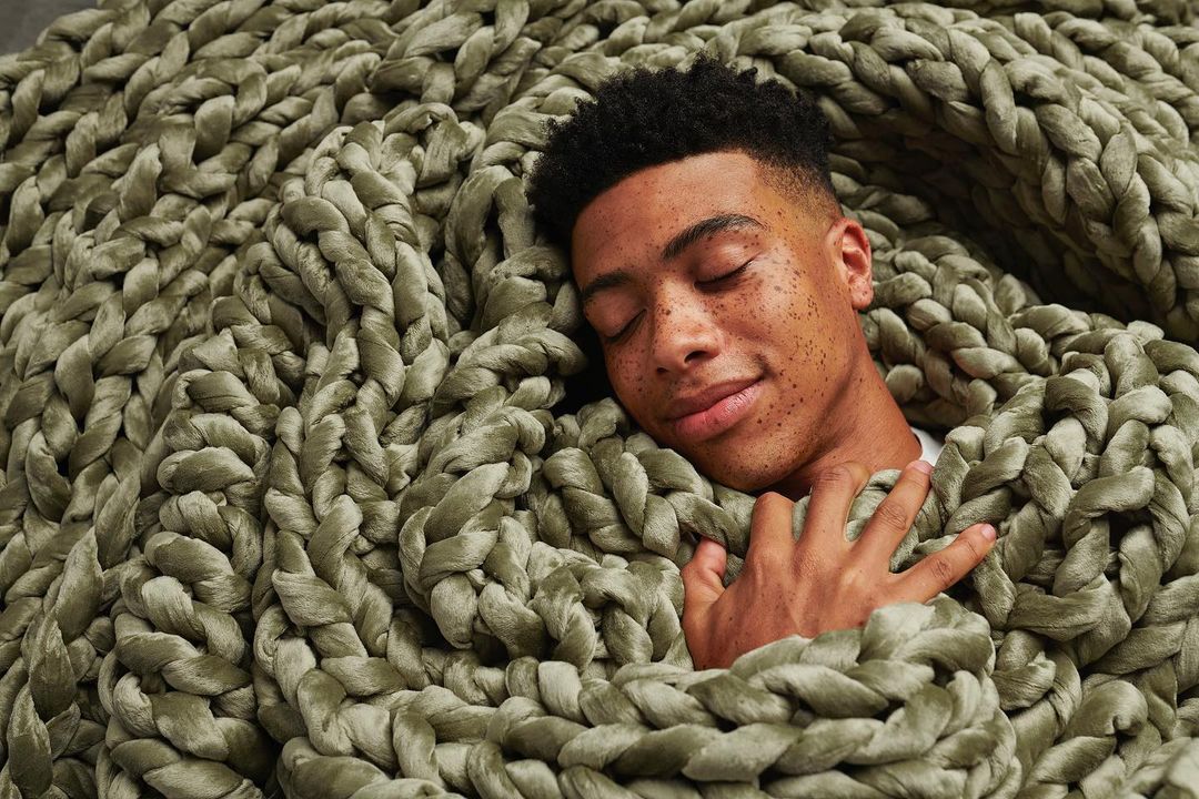 Adult male with dark hair sleeps soundly while nestled underneath a thick, knitted, sage-green blanket in a relaxed position.