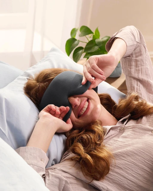 Smiling red-haired woman wearing gray sleep mask lying in bed with blue sheets in brightly lit interior with a green potted plant.