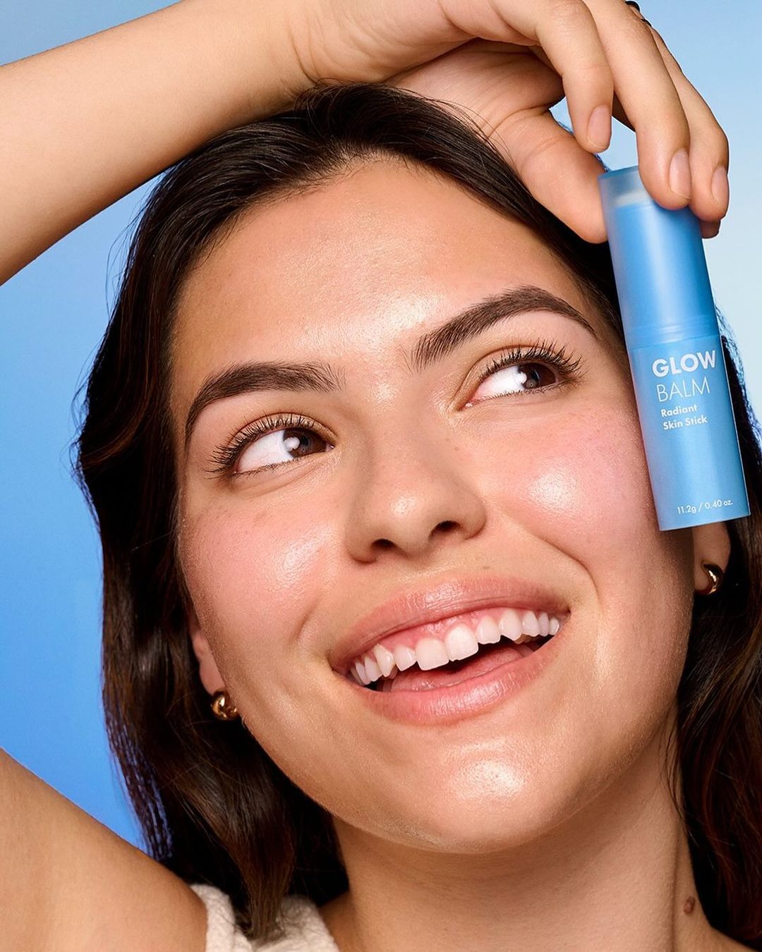 Smiling woman holding a blue tube of Glow Balm skincare product in an eye-level beauty shot against a light blue backdrop.