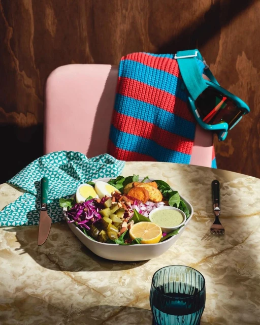 A vibrant salad bowl with eggs, cabbage, potatoes, and lemon, set on a marble table with a glass of water and colorful background.