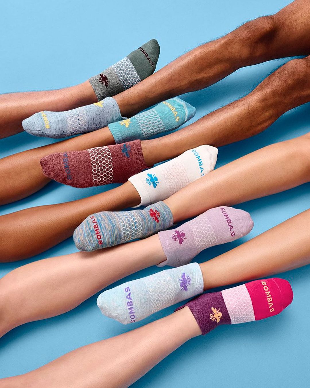 Variety of colorful Bombas socks displayed on multiple human legs against a light blue background, showcasing a range of patterns and designs.