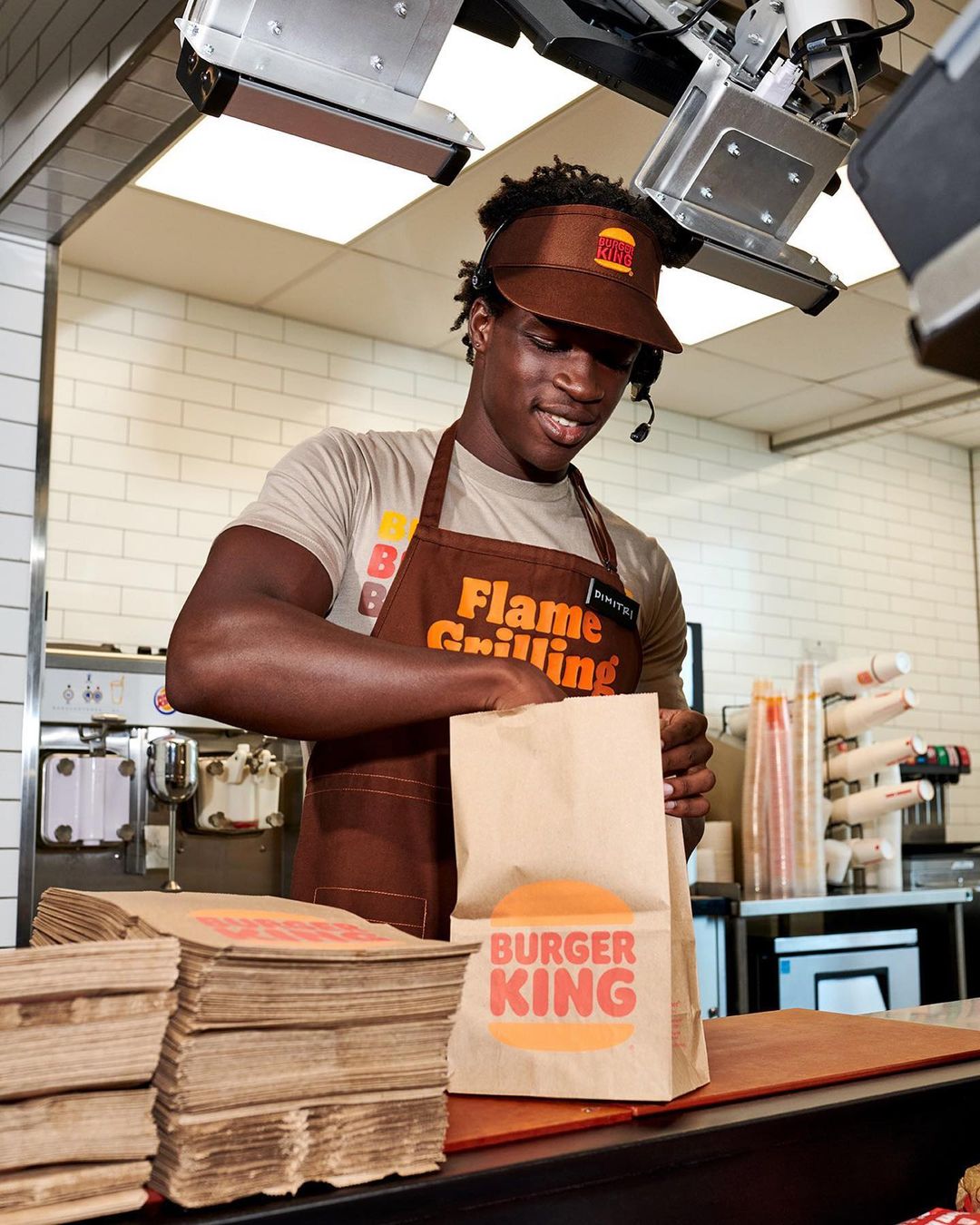Burger King employee prepares a food order at the counter wearing a headset and visor, with stacks of bags and equipment behind him.
