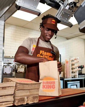 Burger King employee prepares a food order at the counter wearing a headset and visor, with stacks of bags and equipment behind him.
