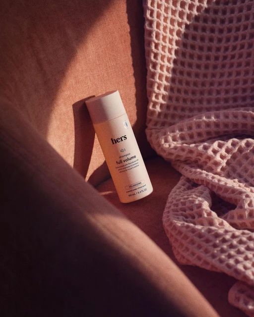 Beige bottle of Hers full volume shampoo rests on peachy-pink textile alongside waffle-patterned pink towel, bathed in warm light.